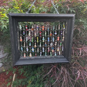 May include: A framed wall hanging with a black, distressed wooden frame. Inside, a wire mesh holds numerous colorful, dangling beads. The beads are various shapes and sizes, with a mix of colors including red, blue, yellow, and clear.