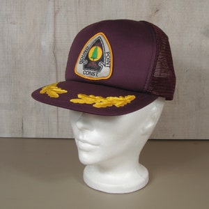 May include: A burgundy trucker hat with a mesh back and a curved brim. The hat features a patch with the text "ROUND POINT CONST" and a tree graphic. Gold leaf embroidery adorns the brim.