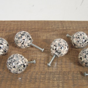 May include: Six round cabinet knobs with a speckled pattern in shades of black, white, and beige. The knobs have a silver metal screw attachment.