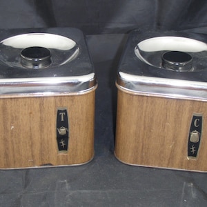 May include: Two vintage metal canisters with woodgrain finish. The canisters have silver lids and are labeled "T" and "C".