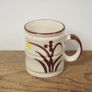 May include: A white ceramic mug with a brown rim and a brown floral design. The design features stylised leaves and a yellow circle.
