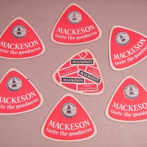 May include: Six red and white triangular cardboard coasters with the text "Mackeson taste the goodness" printed on them. The coasters feature a black and white image of a bottle cap with a lighthouse on top.