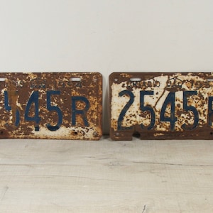 May include: Two vintage Ontario license plates from 1953. The plates are rusty and have blue numbers and letters on a white background. The plates read "2145R" and "2545R".