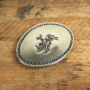 May include: Silver belt buckle with a rope design border and a raised image of a cowboy riding a bucking bull.