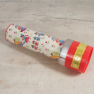 May include: A red and white cardboard tube with a colorful illustration of people and animals. The tube has a silver cap and a red end cap with the words "Musical Spin" in gold lettering.