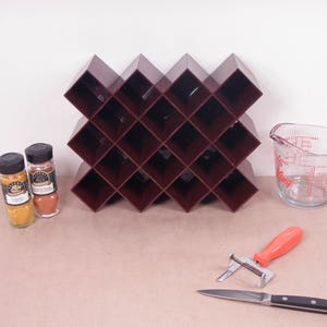 Peut inclure: Un casier à vin marron foncé avec un motif en losange, deux pots d'épices, un verre doseur transparent, un éplucheur et un couteau. Le verre doseur a des marques rouges. Les pots d'épices ont des couvercles noirs.