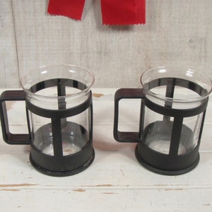 May include: Two clear glass mugs with black plastic handles and a black plastic frame around the glass. The mugs are sitting on a white wooden surface.