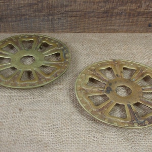 May include: Two antique brass pot holders with a decorative, circular design. The holders have a central circle with eight spokes radiating outwards, each spoke ending in a rounded point. The outer edge of the holders is decorated with a row of small, raised dots.
