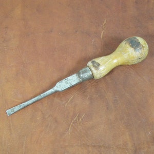 May include: A vintage chisel with a wooden handle and a metal blade. The handle is light yellow with dark spots, and the blade is silver. The tool is designed for woodworking and is set against a brown background.