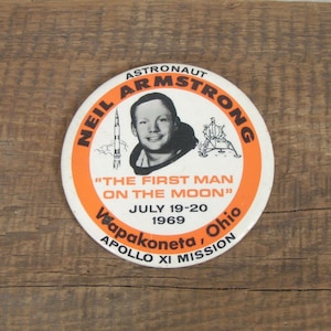 May include: A white button with an orange border featuring a portrait of Neil Armstrong, the text "ASTRONAUT NEIL ARMSTRONG", and the quote "THE FIRST MAN ON THE MOON". The button also includes the text "JULY 19-20 1969 Wapakoneta, Ohio APOLLO XI MISSION".