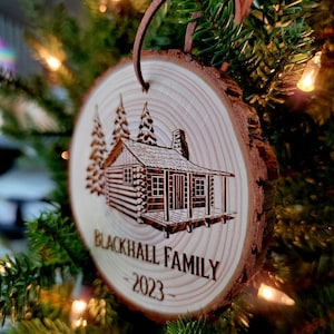 May include: A wooden Christmas ornament shaped like a tree trunk with a cabin and pine trees engraved on it. The ornament is decorated with the text "Blackhall Family - 2023".