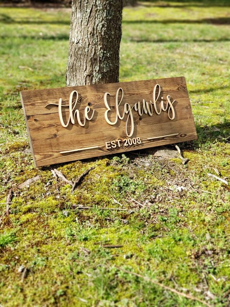 May include: A rustic wooden sign with the text "The Elgawlis Est 2008" in a cursive font. The sign is leaning against a tree trunk and is surrounded by green grass.