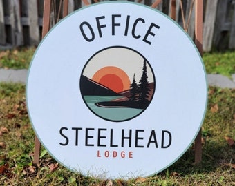 May include: A round, white sign with a blue border. The sign has a logo with a sunset, mountains, and trees. The text "OFFICE" is at the top of the sign and "STEELHEAD LODGE" is at the bottom.