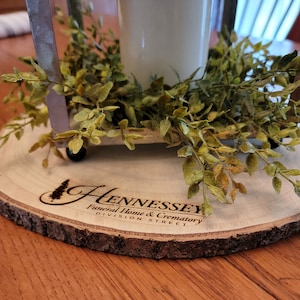 May include: A wooden slice with the text "Hennessey Funeral Home & Crematory Division Street" engraved on it. A small metal lantern with a white candle sits on top of the slice, surrounded by green foliage.