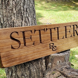 May include: A wooden sign with the word "SETTLER" carved into it. The sign is stained a dark brown color and has a natural wood grain. The letters are in a serif font and are raised from the surface of the sign. The sign is resting on a tree stump.