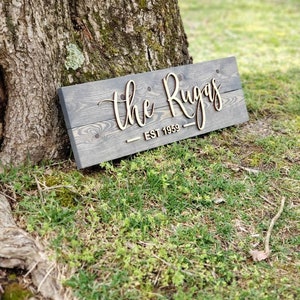May include: A wooden sign with the text "The Rugas - Est 1959" in cursive lettering. The sign is gray with a wood grain texture and the lettering is a light brown color.