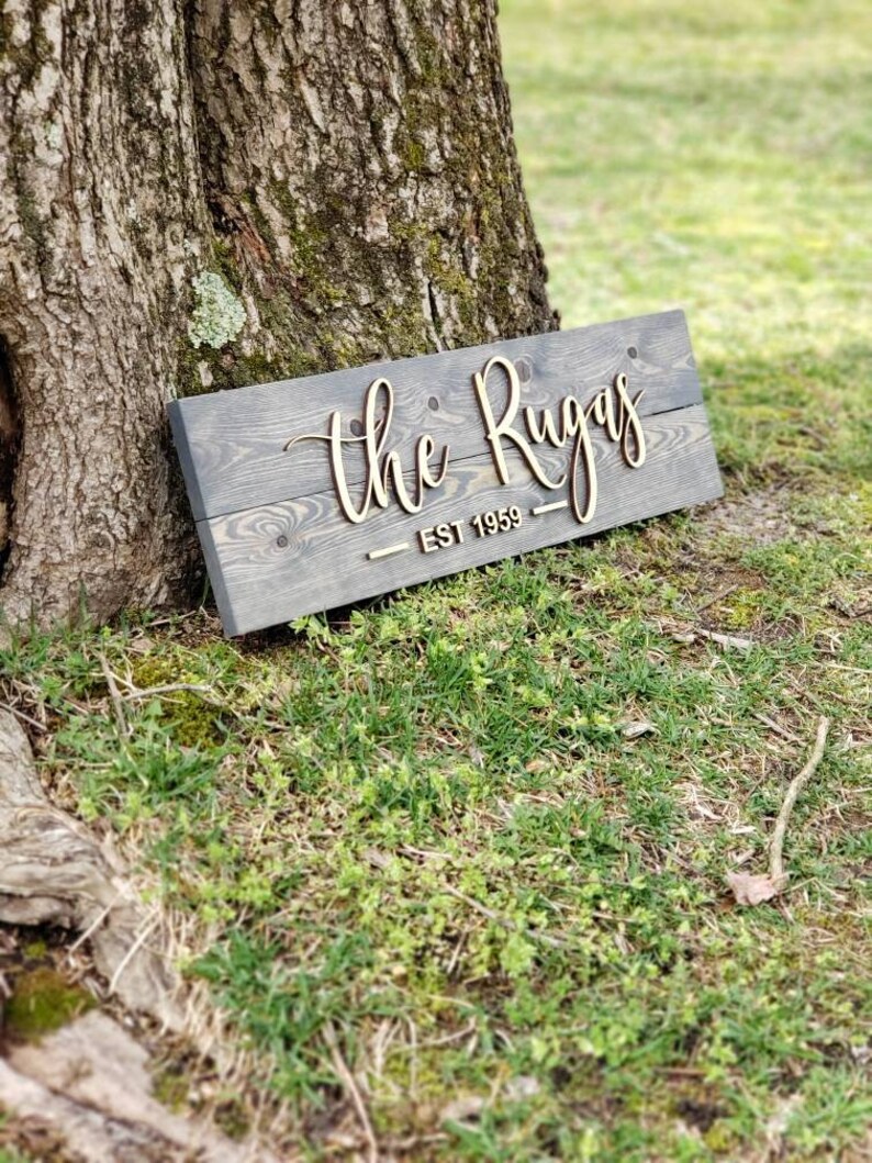 May include: A wooden sign with the text "The Rugas - Est 1959" in cursive lettering. The sign is gray with a wood grain texture and the lettering is a light brown color.