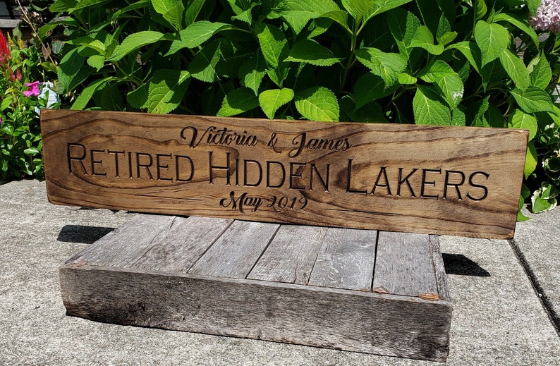 May include: A wooden sign with the text "Victoria & James Retired Hidden Lakers May 2019" engraved on it.