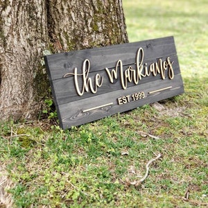 May include: A wooden sign with the text "The Markianos EST 1999" in gold lettering. The sign is leaning against a tree trunk.
