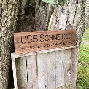 May include: A wooden sign with the text "USS SCHNEIDER - FULL SPEED AHEAD" carved into it. The sign is brown and has a rustic look. It is leaning against a tree trunk.