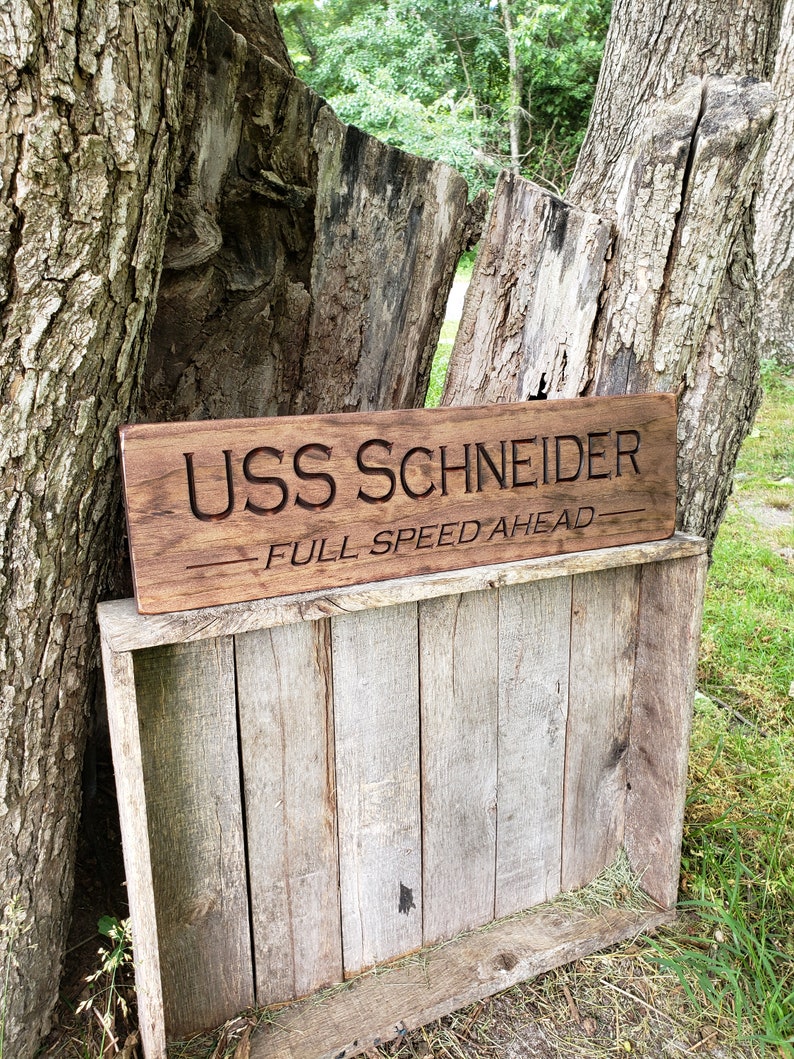 May include: A wooden sign with the text "USS SCHNEIDER - FULL SPEED AHEAD" carved into it. The sign is brown and has a rustic look. It is leaning against a tree trunk.