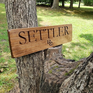 May include: A wooden sign with the word "SETTLER" carved into it. The sign is attached to a tree trunk with a green grassy background.