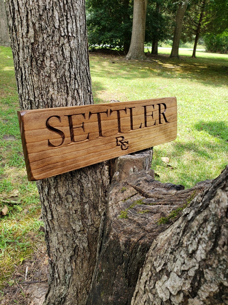 May include: A wooden sign with the word "SETTLER" carved into it. The sign is attached to a tree trunk with a green grassy background.