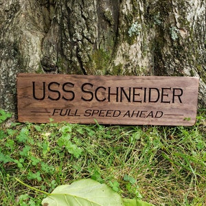 May include: A wooden sign with the text "USS SCHNEIDER - FULL SPEED AHEAD" engraved on it. The sign is brown and has a rustic look.