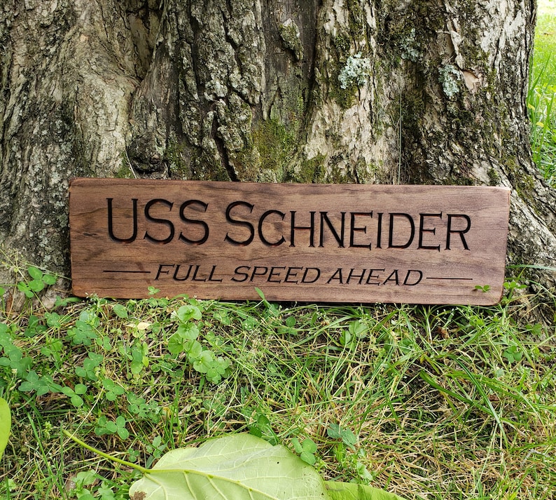 May include: A wooden sign with the text "USS SCHNEIDER - FULL SPEED AHEAD" engraved on it. The sign is brown and has a rustic look.
