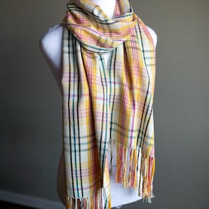 May include: A yellow, pink and green plaid scarf with fringe on the ends. The scarf is draped around a white mannequin.