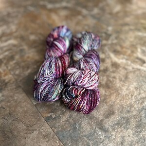 May include: Two skeins of yarn in a purple, white, and black colorway. The yarn is a variegated blend with a subtle shimmer.