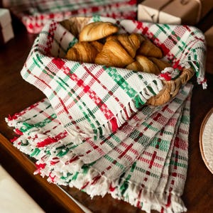 May include: A basket filled with croissants, lined with a red, green and white plaid fabric with fringe. The basket is sitting on a wooden table with other gifts wrapped in white paper with red ribbon.