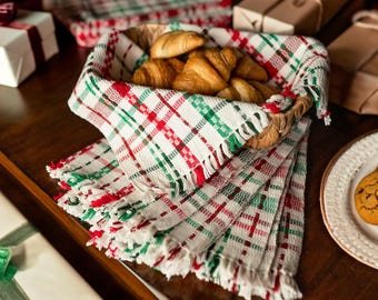 Op de afbeelding: Een mand gevuld met croissants, gevoerd met een rood, groen en wit geruite stof met franjes. De mand staat op een houten tafel met andere geschenken die zijn ingepakt in wit papier met rood lint.