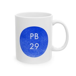 May include: White ceramic mug featuring a blue circle with the white text "PB 29". The mug has a curved handle and is set against a white background.
