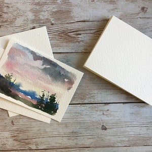 May include: Watercolor landscape art cards with matching envelopes. The cards feature a sunset scene with pink, blue, and gray hues. A stack of blank cards is also visible, ready for use. The cards are on a wooden surface.