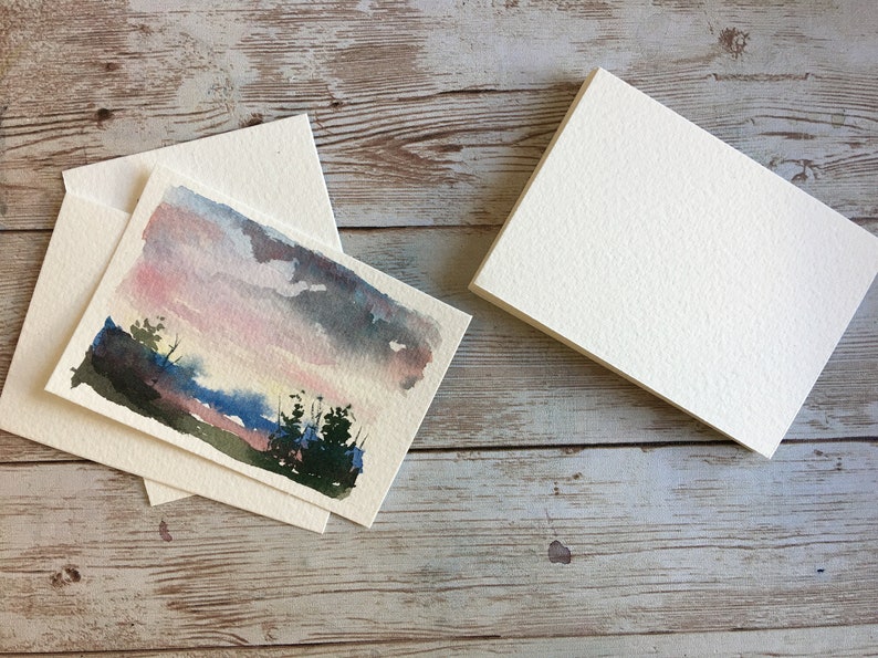 May include: Watercolor landscape art cards with matching envelopes. The cards feature a sunset scene with pink, blue, and gray hues. A stack of blank cards is also visible, ready for use. The cards are on a wooden surface.