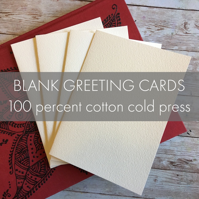 Peut inclure: Un empilement de cartes de v&oelig;ux vierges, de couleur cr&egrave;me, avec une surface textur&eacute;e, pos&eacute; sur un journal rouge avec des motifs d&eacute;coratifs noirs. Le texte de l'image indique "BLANK GREETING CARDS 100 percent cotton cold press."