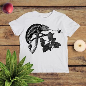 May include: White t-shirt featuring a black and white illustration of a chameleon on a branch with leaves. A bee is flying near the chameleon's tongue.
