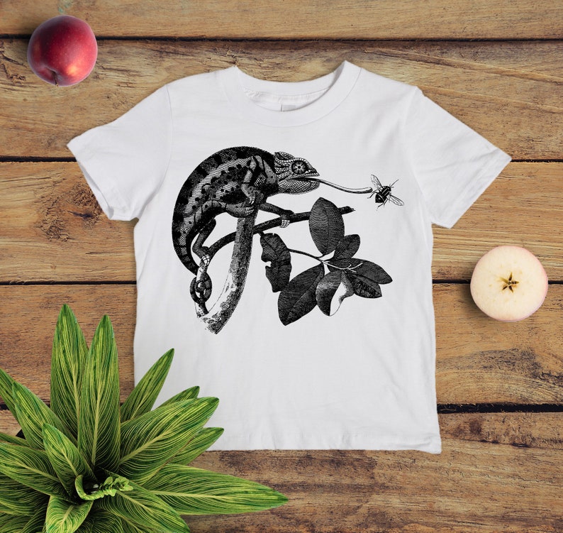 May include: White t-shirt featuring a black and white illustration of a chameleon on a branch with leaves. A bee is flying near the chameleon's tongue.