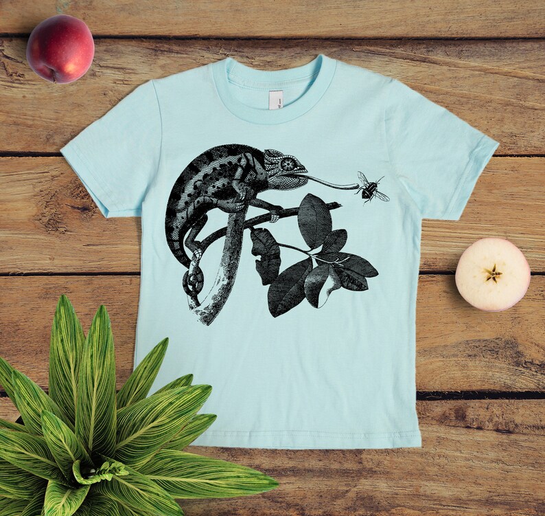 May include: A light blue t-shirt featuring a black and white illustration of a chameleon on a branch with leaves. A small black and white bee is flying near the chameleon's tongue.