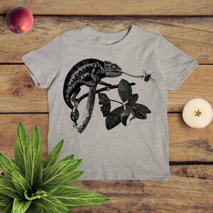 May include: A grey t-shirt featuring a black and white illustration of a chameleon on a branch with leaves. The chameleon is catching a bee with its tongue.
