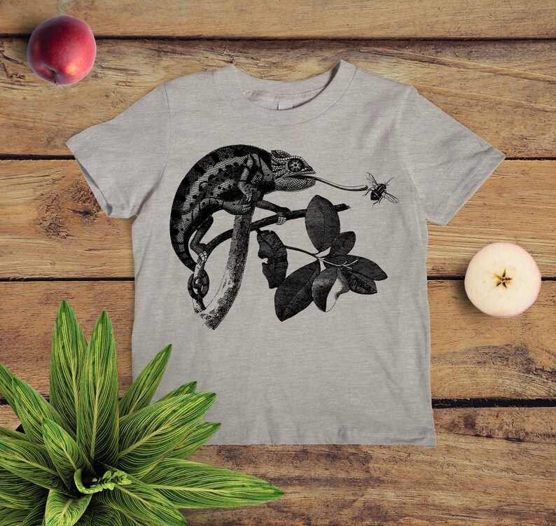 May include: A grey t-shirt featuring a black and white illustration of a chameleon on a branch with leaves. The chameleon is catching a bee with its tongue.