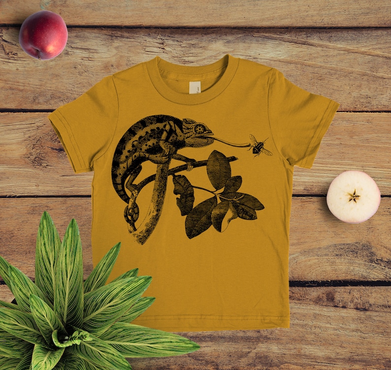 May include: A mustard yellow t-shirt featuring a black and white illustration of a chameleon on a branch with a bee flying nearby.
