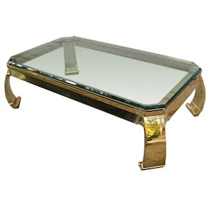 May include: A brass coffee table with a clear glass top and curved legs. The table has a rectangular shape and a modern design.