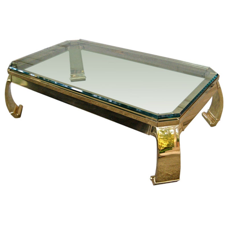 May include: A brass coffee table with a clear glass top and curved legs. The table has a rectangular shape and a modern design.