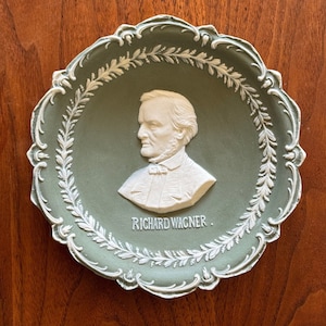 May include: A green ceramic plate with a white raised relief portrait of Richard Wagner. The plate is decorated with a white laurel wreath design.