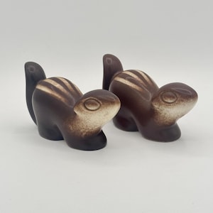 May include: Two brown ceramic squirrel figurines with white stripes. The squirrels are facing forward with their tails curled up.