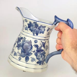 Puede incluir: Una jarra de cerámica blanca y azul con un diseño floral. La jarra tiene un pico curvo y un asa azul. El patrón floral incluye rosas, otras flores y una mariposa. La jarra está sostenida por una persona.