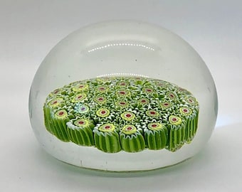 Puede incluir: Un pisapapeles de vidrio transparente con un diseño millefiori verde y blanco. El diseño presenta pequeñas formas de flores con centros rojos.