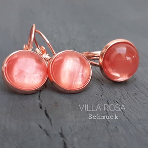 Puede incluir: Un conjunto de joyas con un anillo y pendientes. El anillo y los pendientes tienen un engaste de metal oro rosa con una piedra redonda translúcida de color melocotón. El texto "VILLA ROSA Schmuck" es visible.